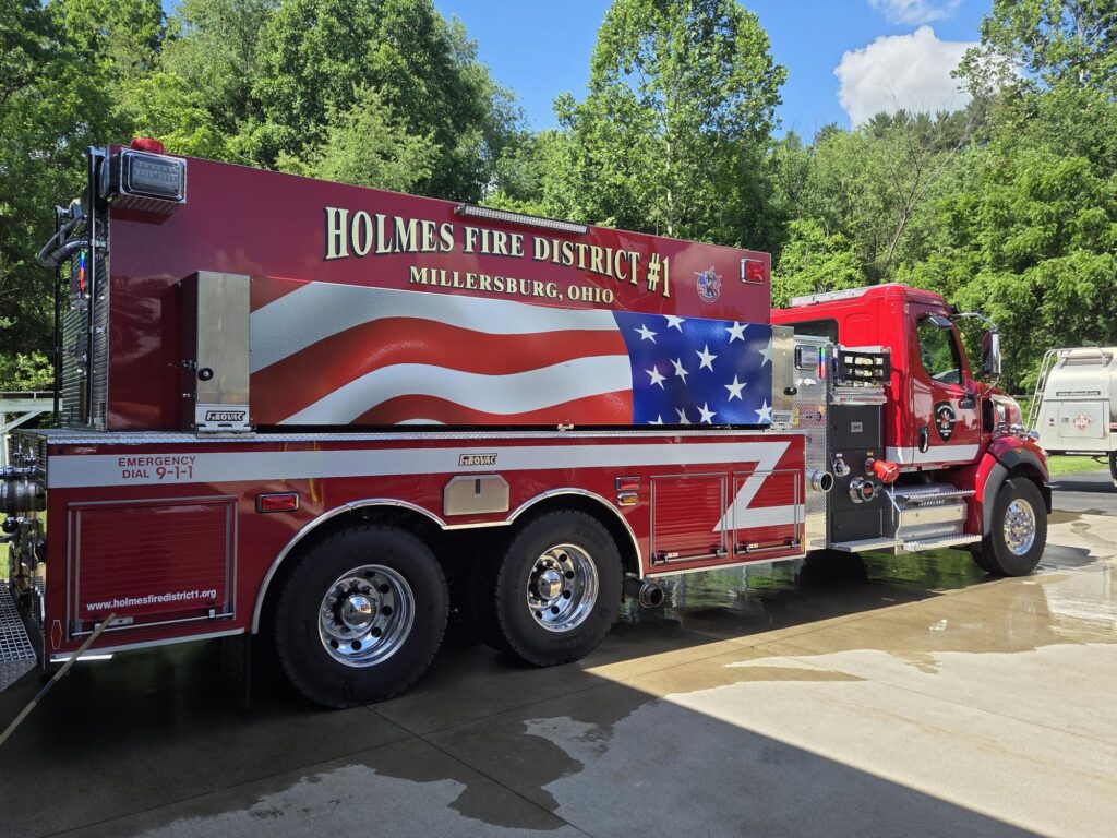 A red fire truck labeled “Holmes Fire District #1 Millersburg, Ohio” features an American flag design on its side, parked on a sunny day with trees and blue sky in the background.