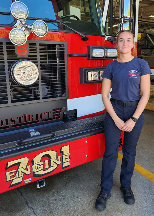 A firefighter is standing in front of a red fire engine with "Engine 701" written on the front. The firefighter is wearing a navy blue uniform, smiling, and posing with hands clasped in front. The background shows part of the fire station interior.
