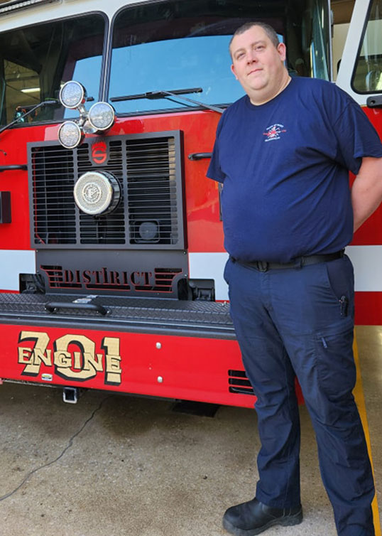 A firefighter in a navy blue uniform stands next to the front of a red fire truck labeled "Engine 701." The firefighter has short hair and is smiling, with folded arms resting on the fire truck's grill. The vehicle and station garage are in the background.