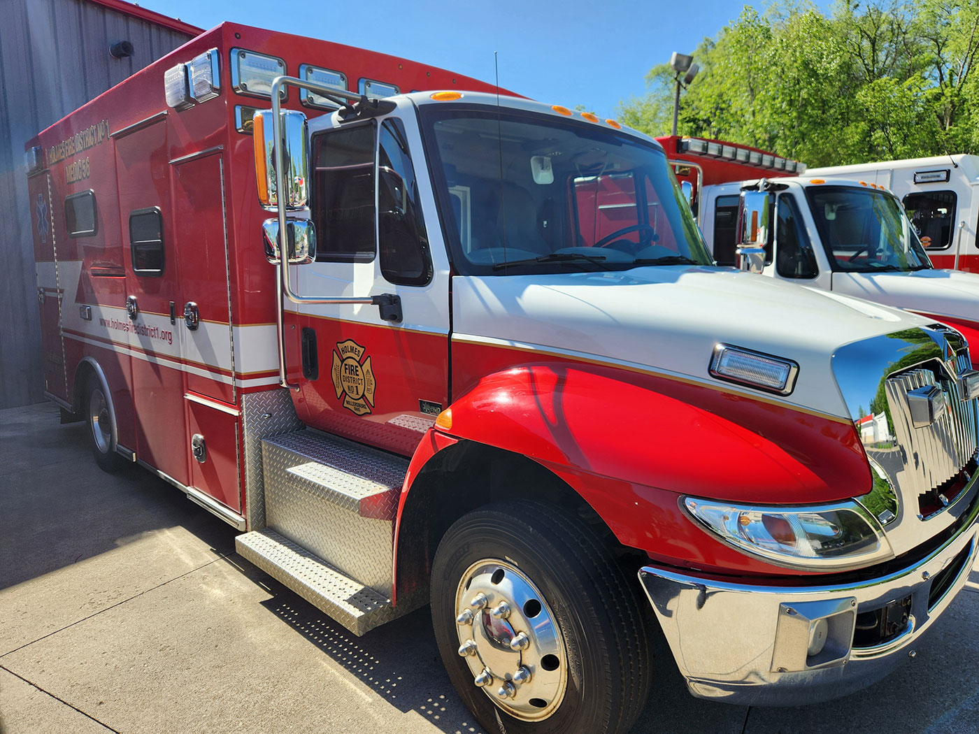 A bright red and white emergency medical services truck is parked outside a building with another emergency vehicle visible in the background. The truck features a reflective emblem and has a side compartment and steps for easy access. Trees are in the background.