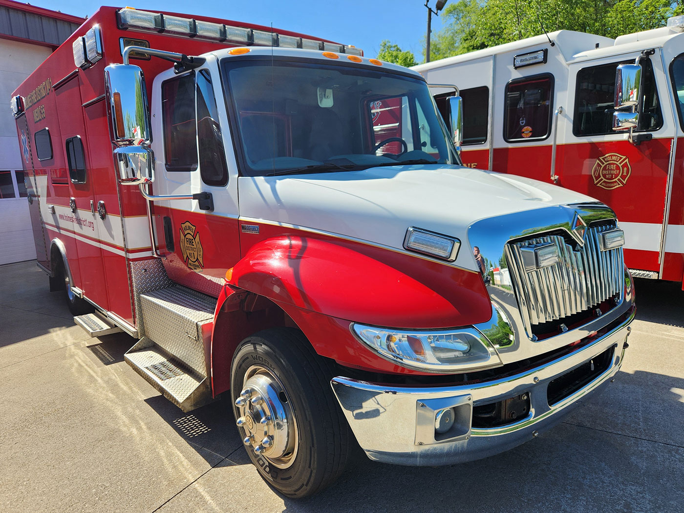 A bright red ambulance is parked outdoors near a fire truck. The vehicle has a white stripe along its side, emergency lights on the roof, and a reflective chrome grille. An emblem is visible on the door. The background shows a building and trees.