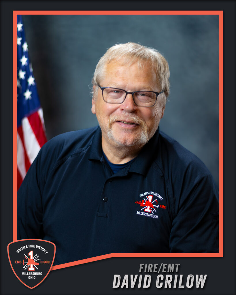 Smiling man with glasses and gray hair in a navy Holmes Fire District shirt, seated in front of a US flag and gray background. Fire/EMT badge and name “David Crilow” displayed at the bottom.