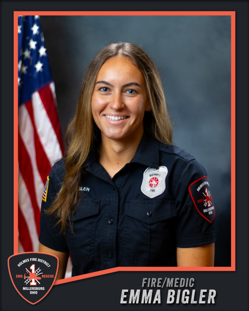 A woman in a black fire department uniform smiles in front of an American flag and gray backdrop. Text on the image reads “Fire/Medic Emma Bigler, Holmes Fire District, Millersburg, Ohio.”.