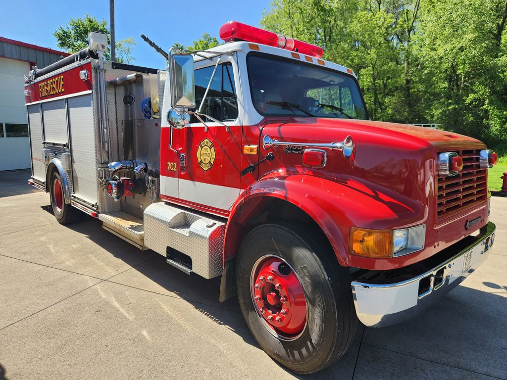 A red and white fire truck is parked outside a fire station on a sunny day. The truck has various equipment and tools stored in compartments, and its polished exterior reflects the sunlight. Green trees and blue sky are visible in the background.
