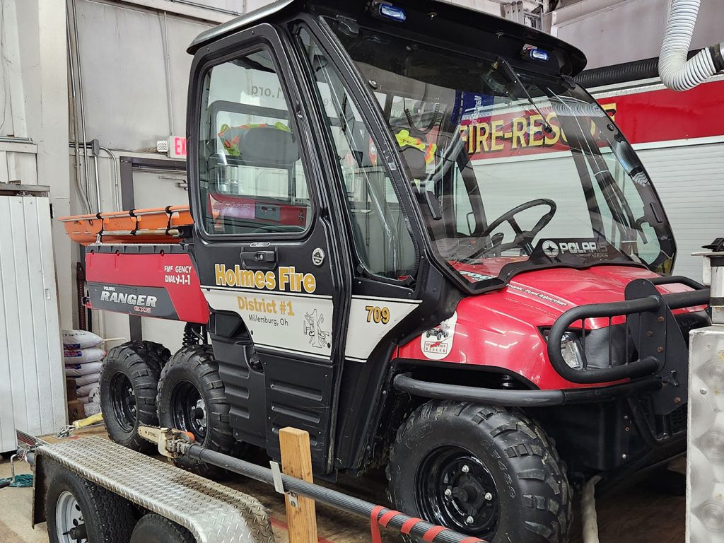 A red and black fire rescue utility vehicle labeled "Holmes Fire District #1" is pictured inside a fire station. The vehicle is equipped with emergency lights and is secured on a trailer, ready for deployment. Various firefighting equipment can be seen in the background.