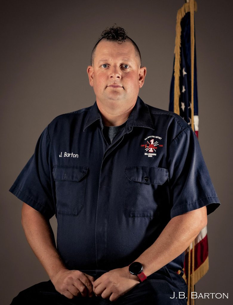 A person in a dark blue uniform with a name tag "J. Barton" sits against a grey background with an American flag partially visible behind them. The person has a serious expression and is wearing a smartwatch on their left wrist. The text "J.B. BARTON" appears in the bottom right corner.