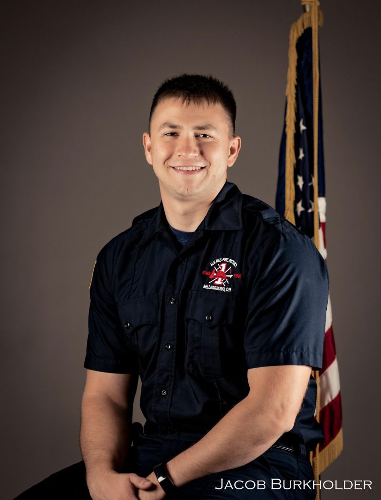 A man in a navy blue uniform is sitting and smiling. He has short hair and is positioned in front of a U.S. flag. The name "JACOB BURKHOLDER" is written at the bottom right of the image.