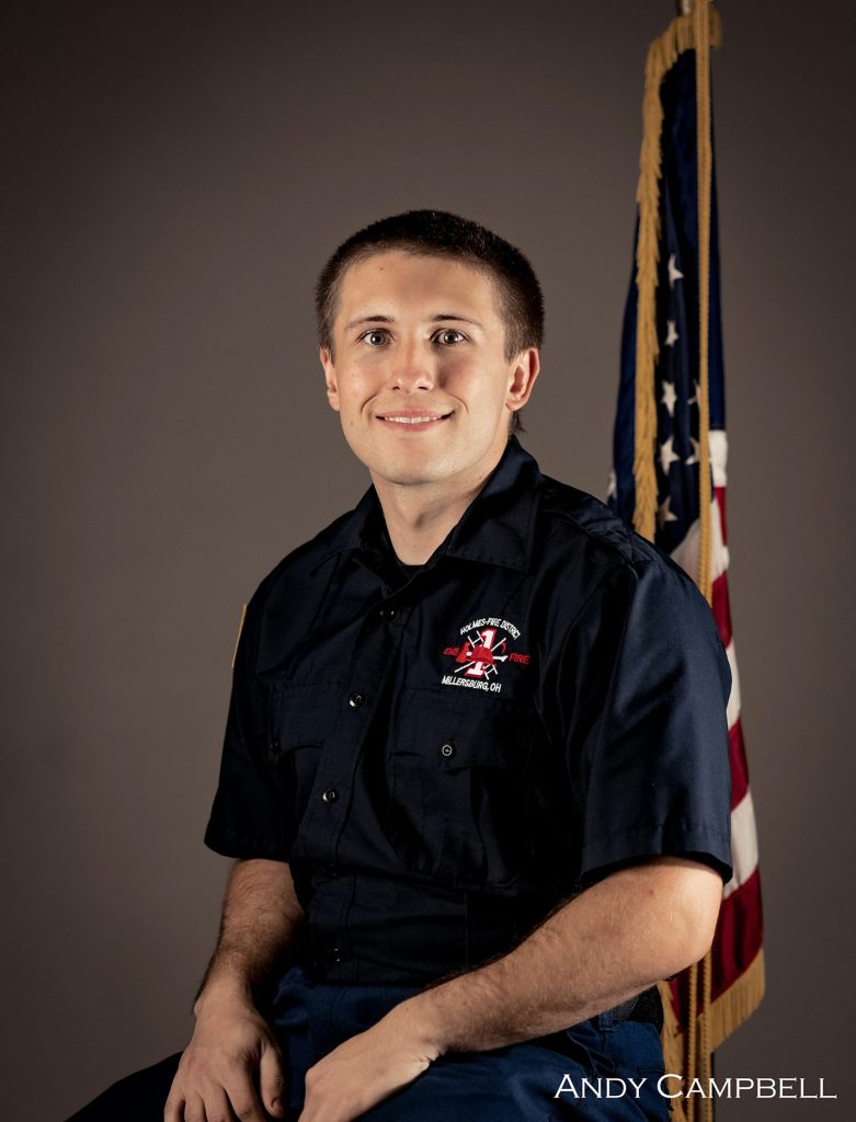 A young adult man with short hair smiles while sitting and wearing a dark uniform embroidered with "Fire Dept" and an emblem on the chest. An American flag stands in the background. The name "Andy Campbell" is visible at the bottom right of the image.