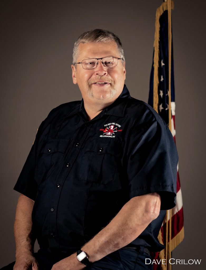 A middle-aged man with gray hair and glasses is dressed in a navy blue uniform, sitting against a neutral background with an American flag beside him. The uniform features a fire department insignia on the chest. The photograph is credited to Dave Crilow.