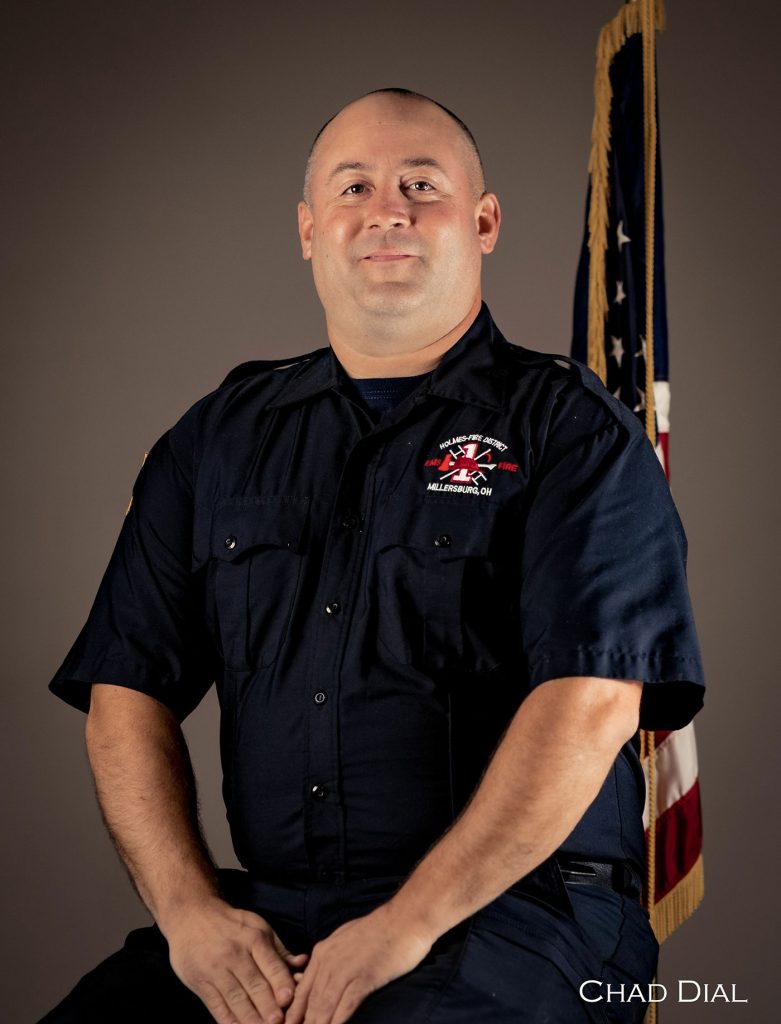 A man in a dark uniform with a firefighter emblem sits in front of an American flag. He has short hair and is smiling slightly. The background is plain, and "CHAD DIAL" is written in the bottom right corner.