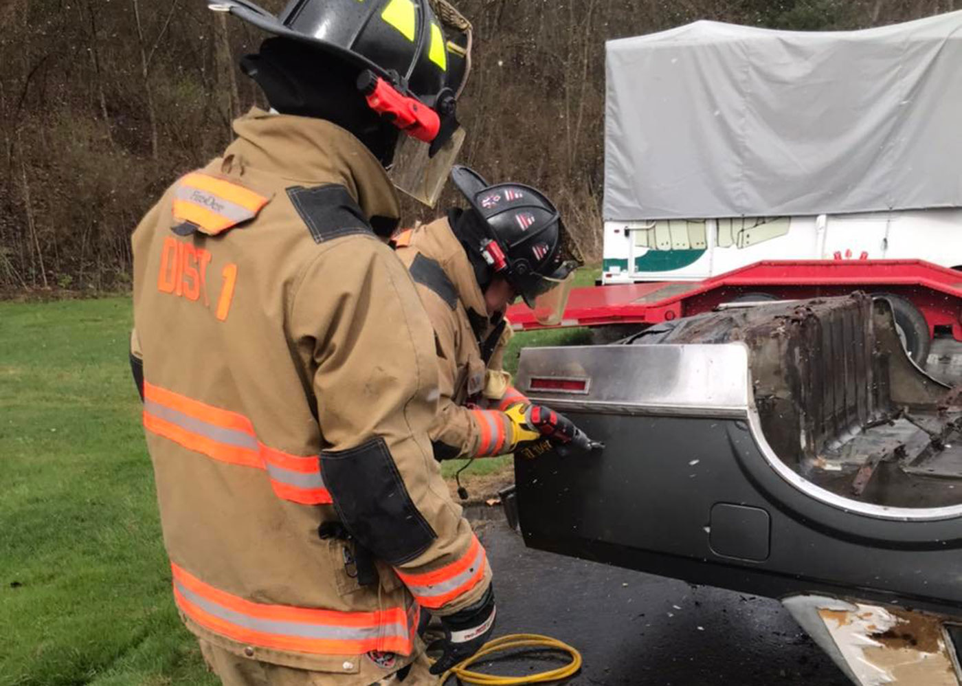 Two firefighters in full gear are using tools to cut into a car's metal frame. One firefighter holds a rotary saw while the other assists. The car appears to be a training vehicle, with other equipment and a covered object visible in the background.