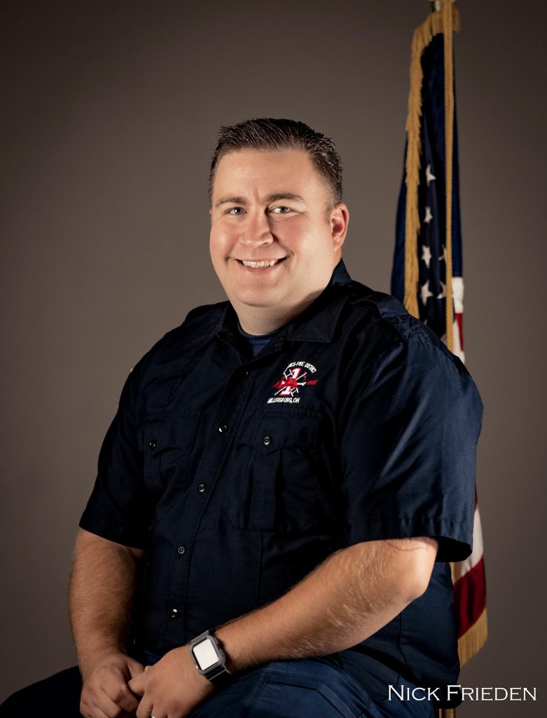 A smiling person with short hair, wearing a dark blue uniform with an embroidered emblem, sits in front of an American flag. The background is a plain gray, and the name "Nick Frieden" is visible in the lower right corner.