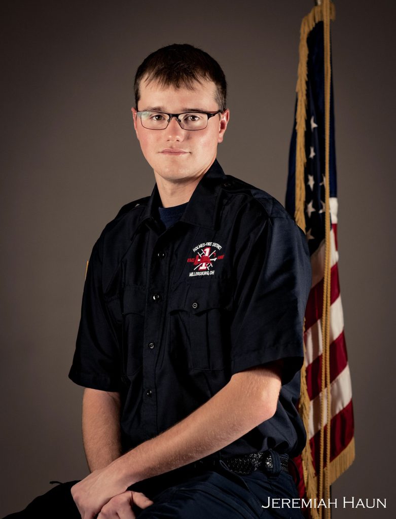 A young man in a dark uniform with a fire department insignia sits confidently in front of an American flag. He has short hair and wears glasses. The background is plain and neutral. The photo is credited to Jeremiah Haun, as indicated in the lower right corner.