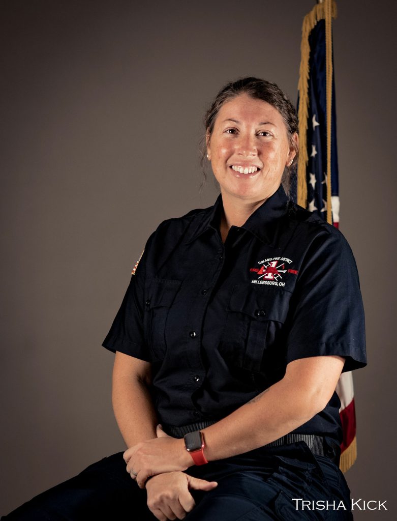 A woman in a dark uniform sits smiling with her hands on her lap. An American flag is partially visible in the background. The uniform displays a patch with a crossed axe and ladder, and text that reads "Malabar, OH." The name "Trisha Kick" is visible in the corner.