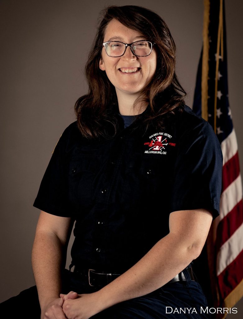 A smiling woman with glasses and long brown hair sits in front of an American flag, wearing a dark uniform with the logo "Medina Fire Department" and “Medina, OH” on it. The name "Danya Morris" is visible in the bottom right corner.