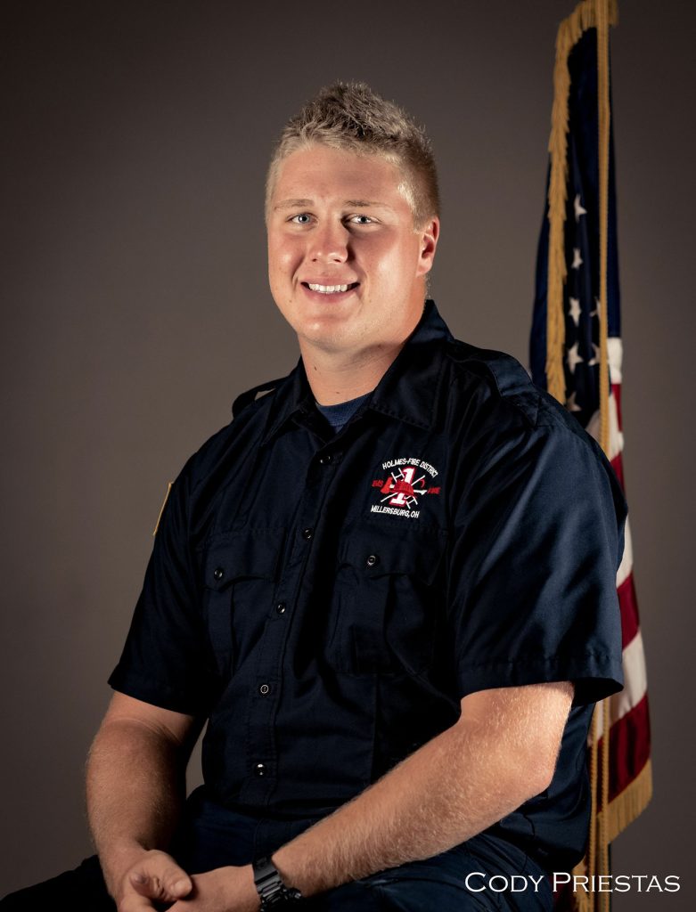 A person in a dark uniform with a fire department emblem sits in front of an American flag, smiling. The individual has short, light-colored hair. The name "Cody Priestas" is visible in text in the bottom right corner.