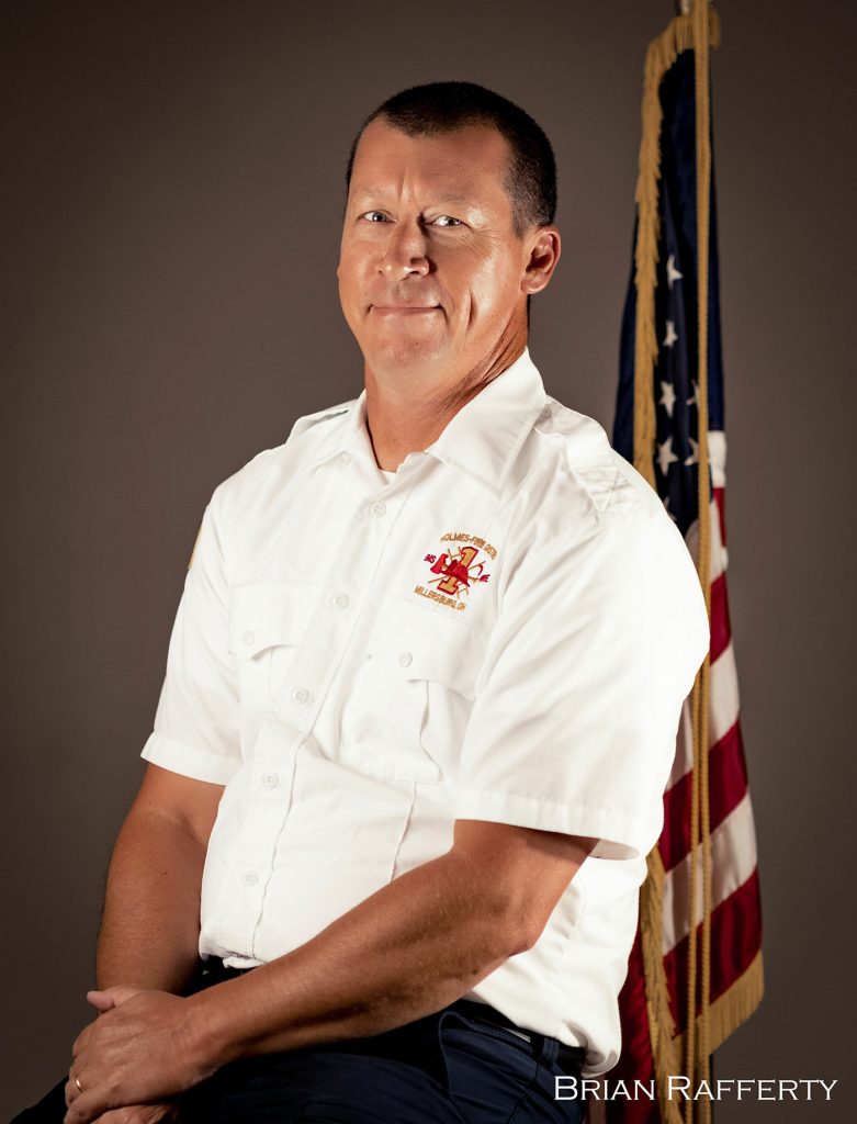 A man in a white uniform shirt with a fire department logo sits against a plain background with the American flag behind him. He has short, dark hair and a slight smile. The name "Brian Raffertry" is printed at the bottom right of the image.
