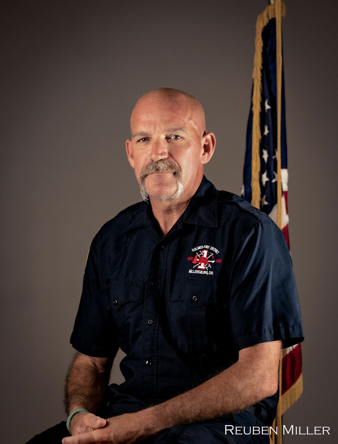 A man with a mustache and shaved head, wearing a dark blue uniform with a fire department logo, sits in front of a gray background. An American flag is behind him. The name "REUBEN MILLER" is printed in white at the bottom right corner of the image.