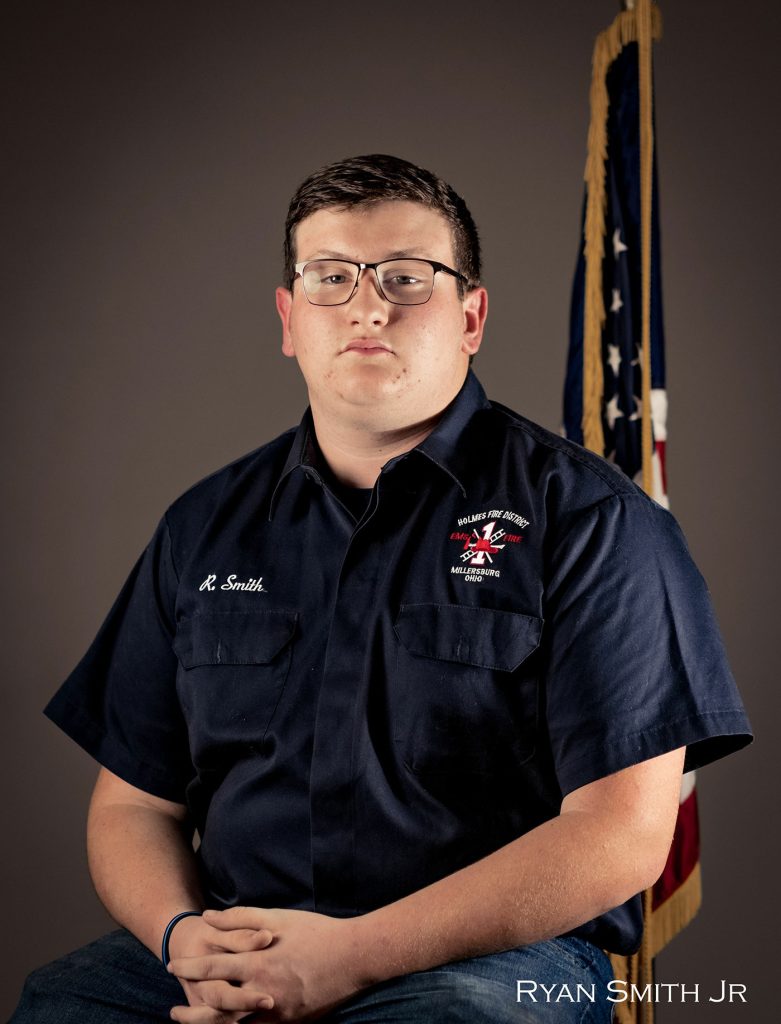 A person wearing glasses and a dark blue shirt with a fire department emblem sits in front of a neutral background. The shirt has a name tag that reads "R. Smith." An American flag is partially visible in the background. Text in the corner reads "RYAN SMITH JR.