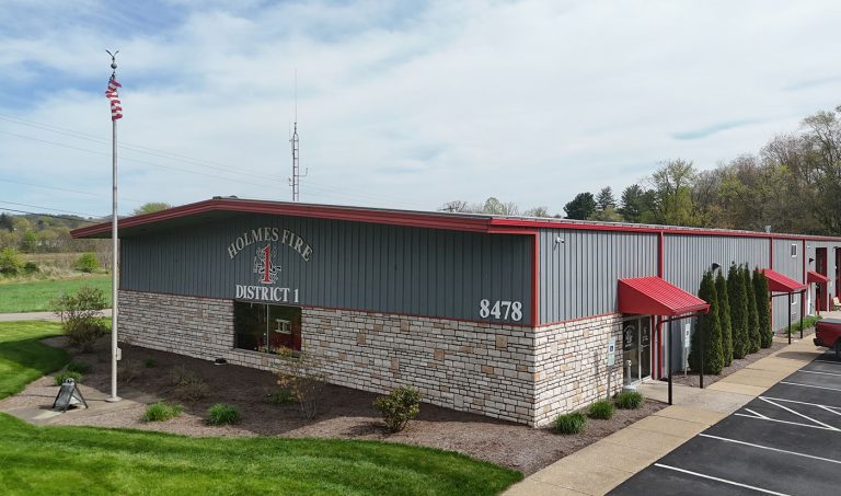 A one-story fire station building with gray metal siding, red trim, and a sloped roof. The building is labeled "Holmes Fire District 1" and has the address number "8478" displayed. An American flag flies on a pole in front, surrounded by a well-maintained lawn and bushes.