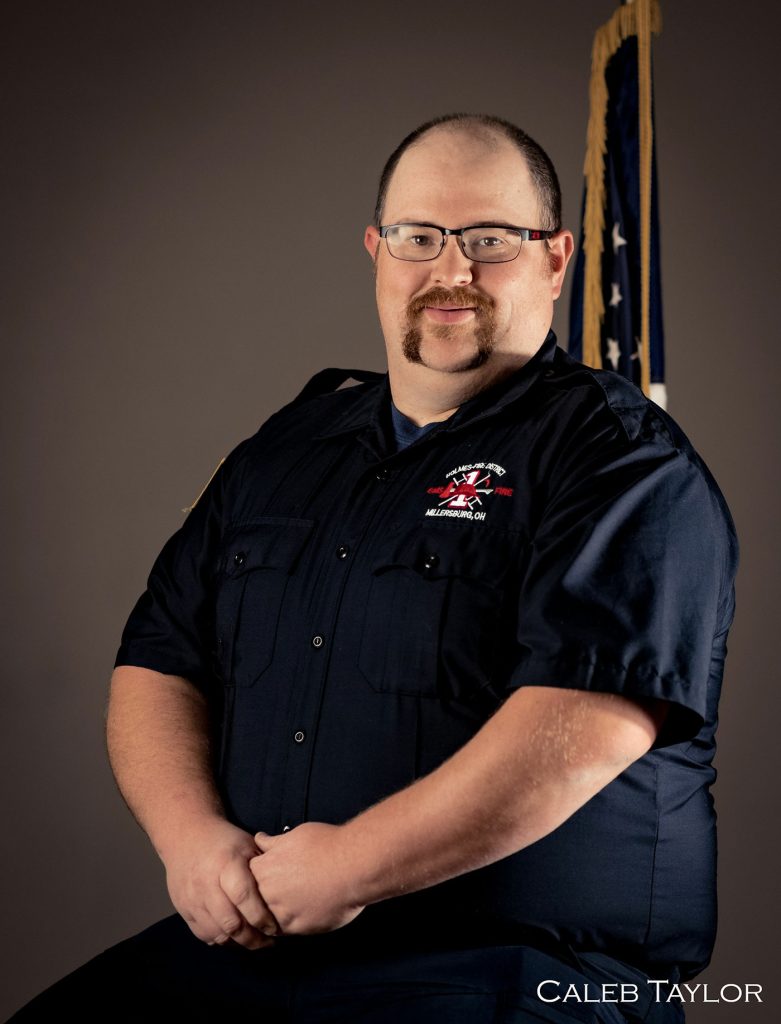 A man wearing glasses and a dark firefighter uniform is posing in front of a plain background with part of an American flag visible. His hands are clasped in front of him and he has a mustache and goatee. The name "Caleb Taylor" is written in the lower right corner.