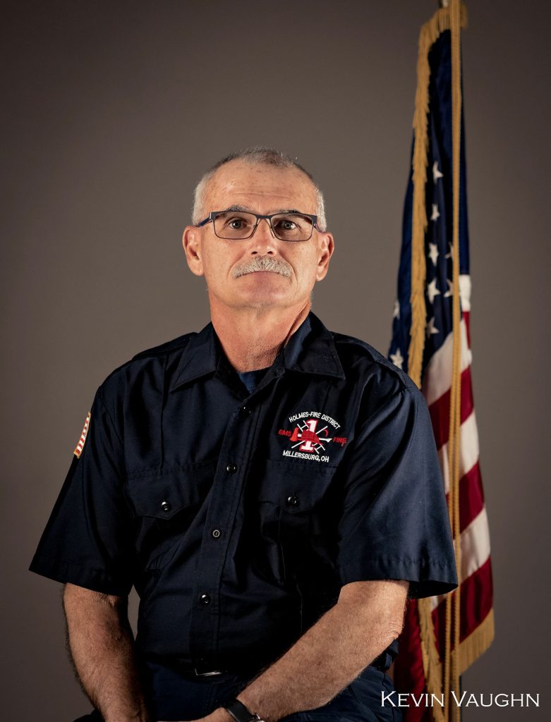 A man with short gray hair, glasses, and a mustache is seated in front of an American flag. He is wearing a navy blue uniform with a patch that includes an American flag and a fire department emblem. The name Kevin Vaughn is printed at the bottom right.