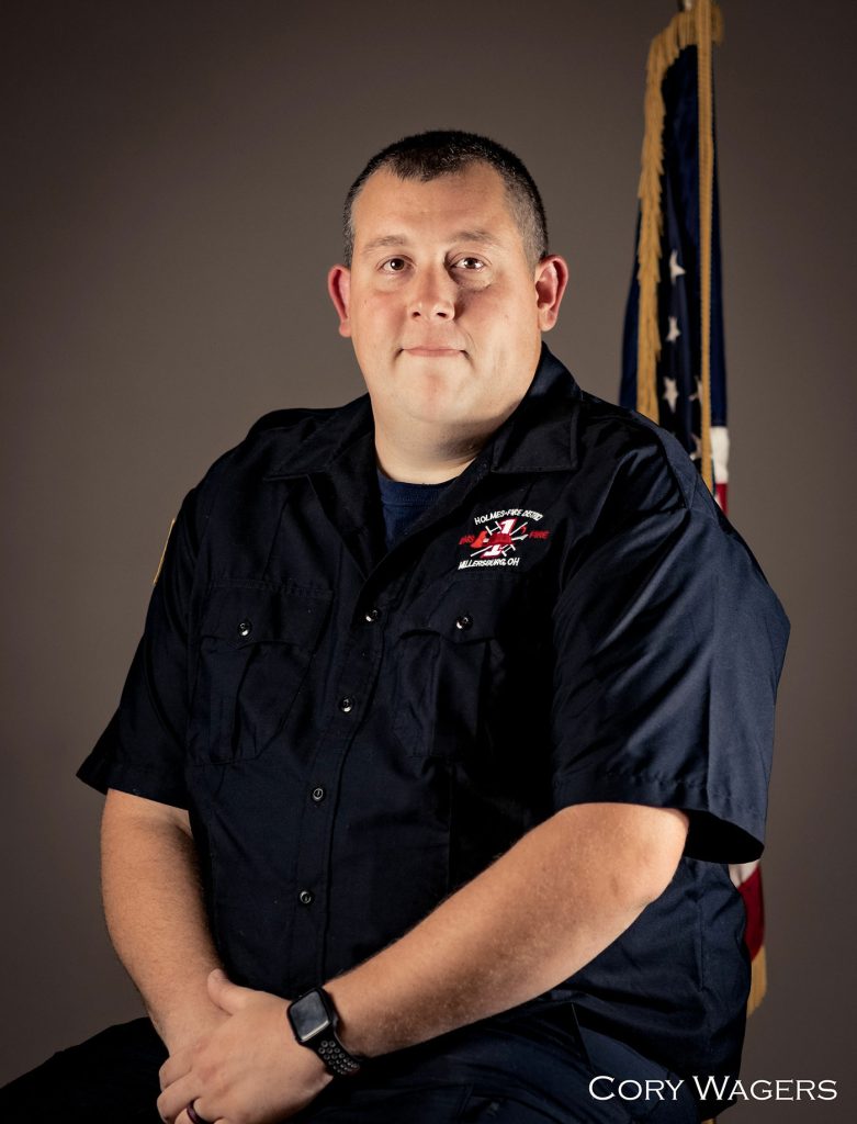 A man in a dark work shirt with an embroidered patch sits for a formal portrait. He has short hair and wears a wristwatch. Behind him is an American flag. The name "Cory Wagers" is visible in the bottom right corner of the image.