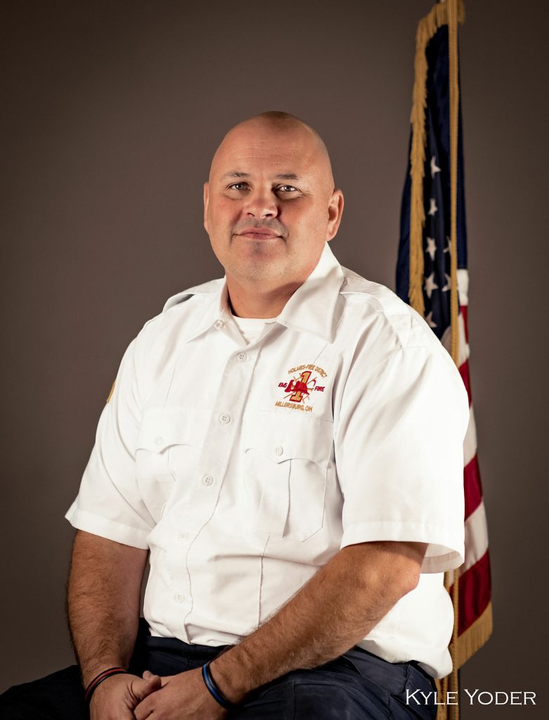A man in a white uniform shirt with a fire department logo sits in front of a flag. He is bald and smiling. In the lower right corner, the text "Kyle Yoder" is visible.