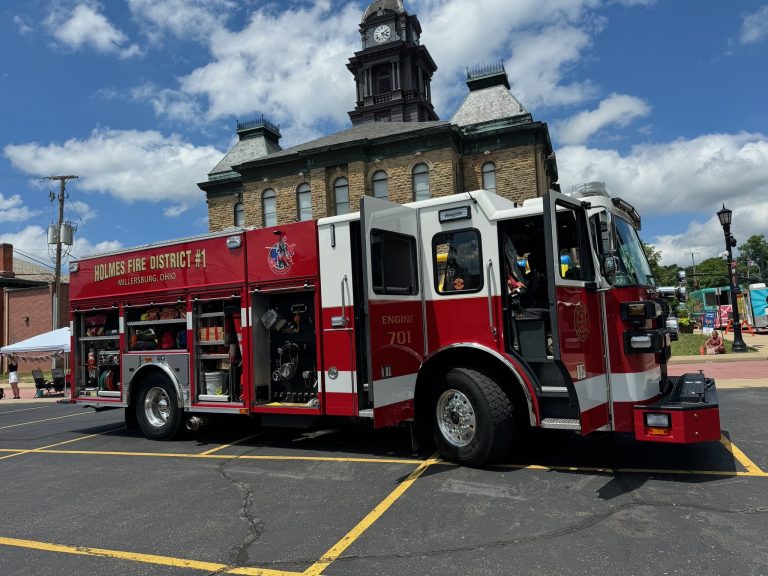 A red fire truck labeled "Holmes Fire District #1, Millersburg, Ohio" parked in front of a historic building with a clock tower. The sky is partly cloudy, and the scene is set in a town square with a few people in the background.