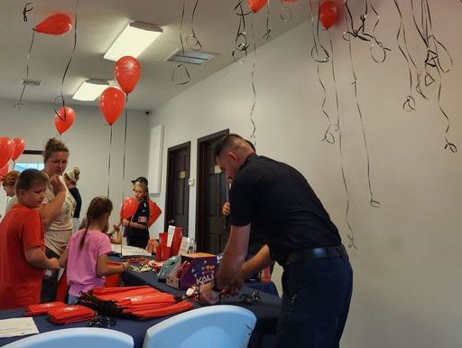 People gather in a room filled with red balloons hanging from the ceiling. A man is preparing items on a table, while children and adults look on. The atmosphere seems festive and organized.