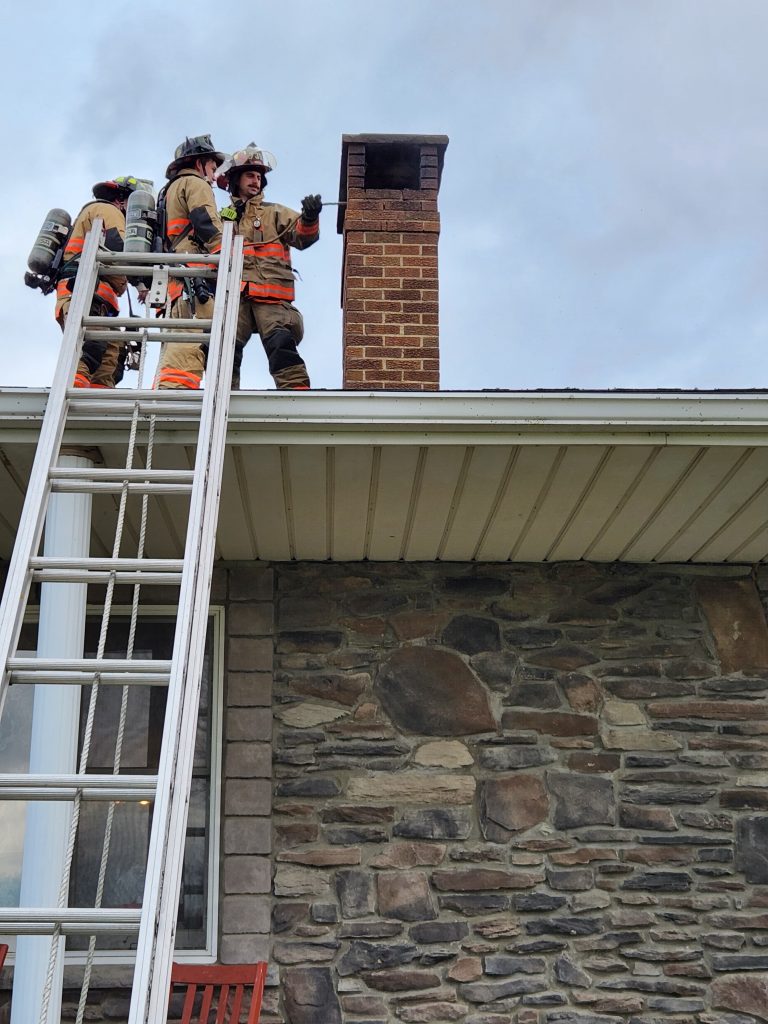 Three firefighters in gear stand on a roof next to a brick chimney, with a tall ladder leaning against the building. They appear to be inspecting the chimney. The house has stonework on its exterior wall. The sky is overcast.