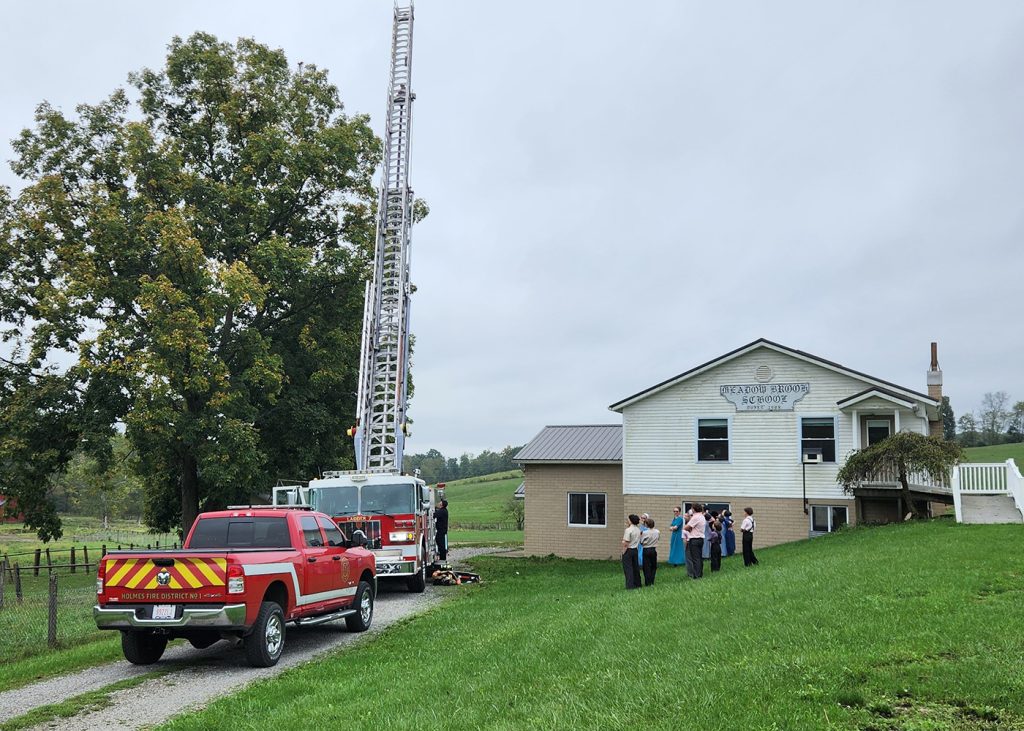 A group of people, including firefighters, stand near a white building with a sign "Branch 309 Legion." A fire truck with an extended ladder is on the grassy lawn, and a red response vehicle is parked beside it. A large tree is nearby.