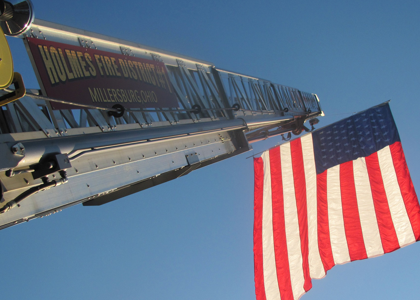 A large American flag is hoisted on a fire truck ladder with "Holmes Fire District #1, Millersburg, Ohio" printed on it. The ladder extends into a clear blue sky.