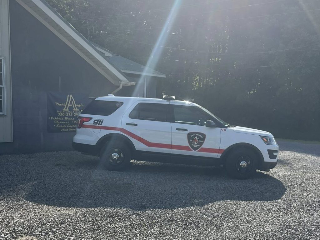 A white police SUV with red stripes and "911" parked on gravel next to a building on a sunny day, with sunlight streaming down and trees visible in the background.