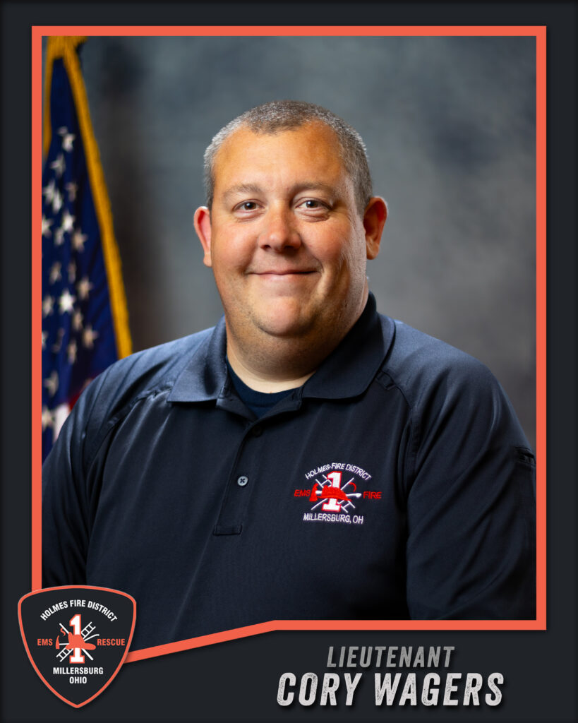 A man in a Holmes Fire District polo shirt smiles in front of a U.S. flag. The text reads: “Lieutenant Cory Wagers,” with the Holmes Fire District logo in the corner.